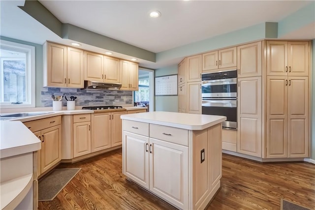 kitchen with light brown cabinetry, under cabinet range hood, wood finished floors, gas stovetop, and double oven