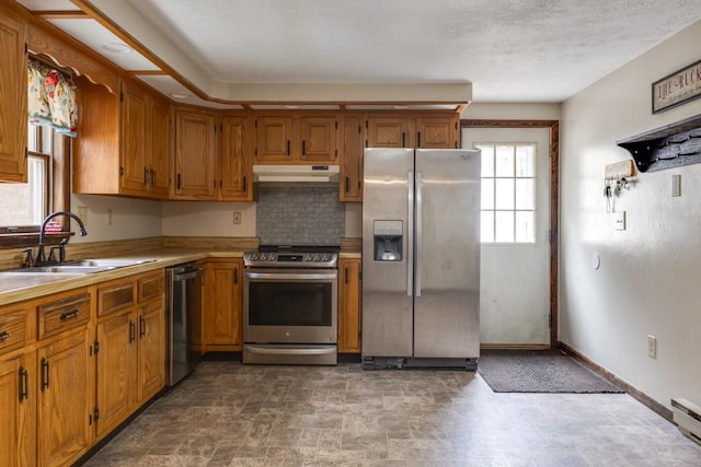 kitchen featuring brown cabinetry, a sink, light countertops, under cabinet range hood, and appliances with stainless steel finishes