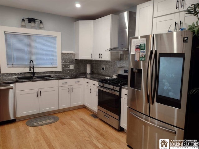 kitchen featuring light wood-style flooring, a sink, stainless steel appliances, dark countertops, and wall chimney exhaust hood