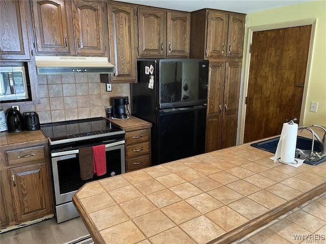 kitchen featuring under cabinet range hood, backsplash, stainless steel electric stove, freestanding refrigerator, and tile counters