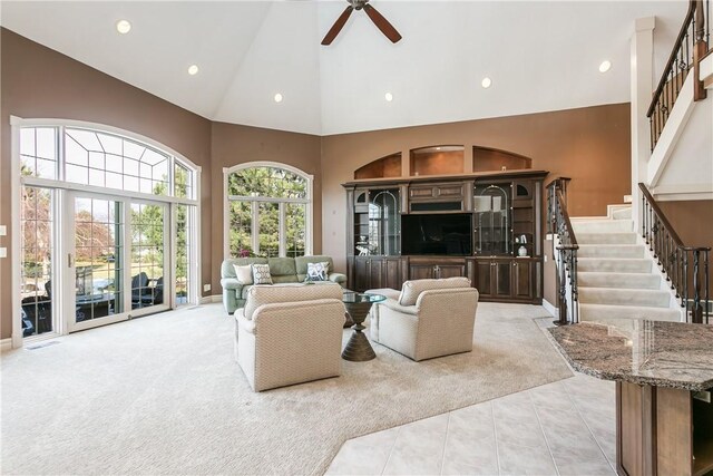 living area with ceiling fan, stairway, light colored carpet, light tile patterned flooring, and high vaulted ceiling