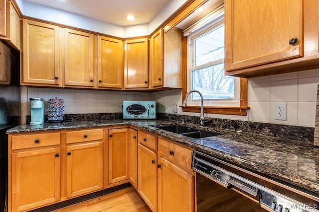 kitchen featuring light wood finished floors, dishwasher, decorative backsplash, dark stone countertops, and a sink