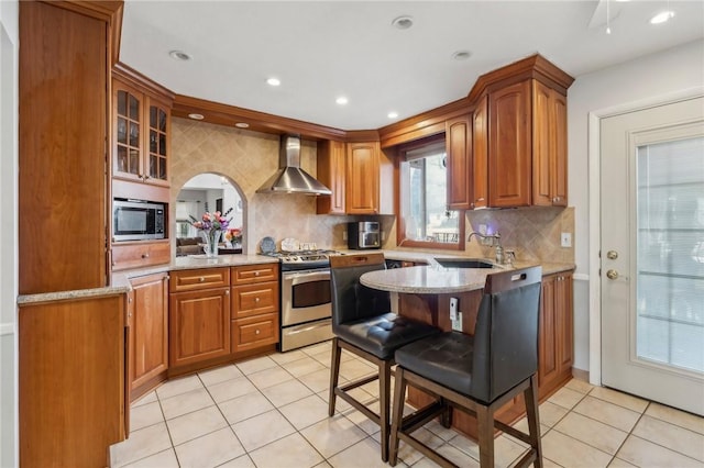 kitchen with light stone countertops, brown cabinetry, stainless steel appliances, wall chimney exhaust hood, and a sink