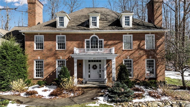 colonial home featuring a balcony, brick siding, roof with shingles, and a chimney