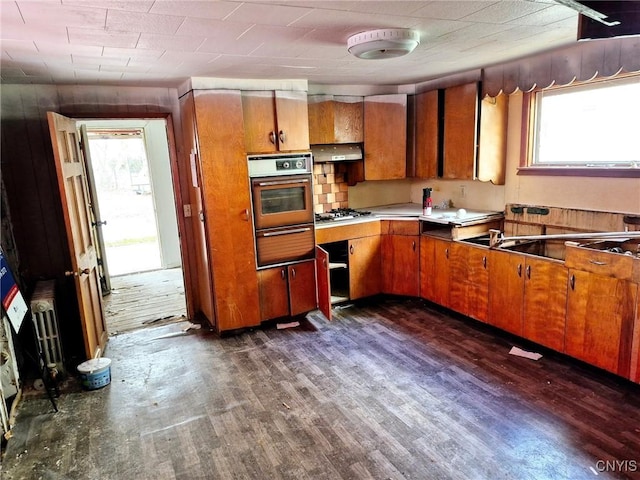 kitchen with dark wood-style flooring, oven, stainless steel gas stovetop, a warming drawer, and brown cabinets