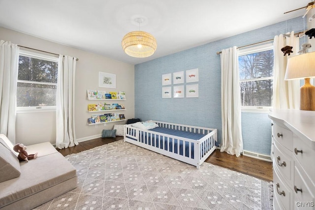 bedroom featuring wood finished floors, baseboards, wallpapered walls, a crib, and an accent wall