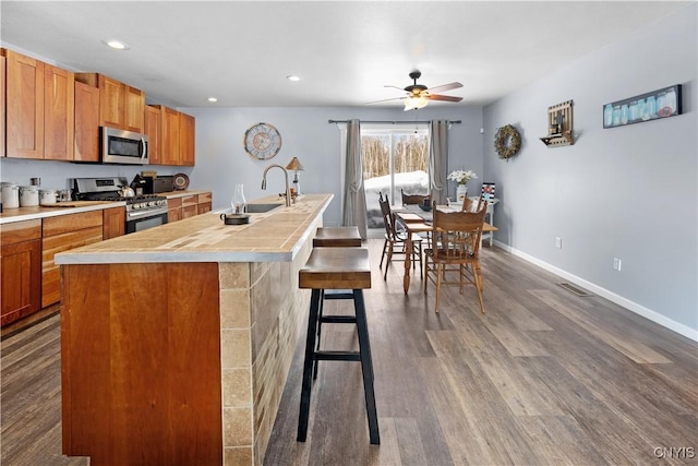 kitchen with baseboards, dark wood finished floors, an island with sink, a sink, and appliances with stainless steel finishes