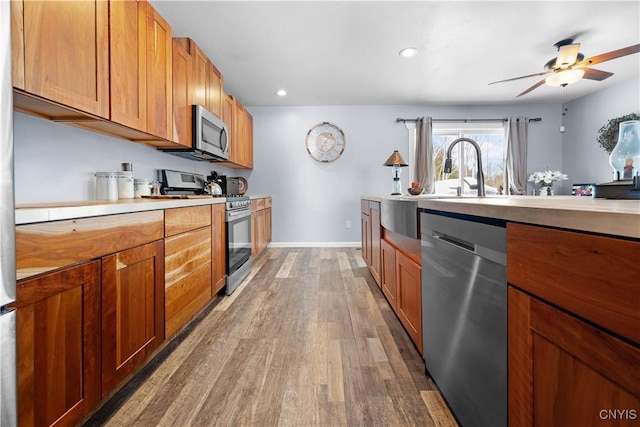 kitchen featuring wood finished floors, appliances with stainless steel finishes, brown cabinetry, light countertops, and baseboards