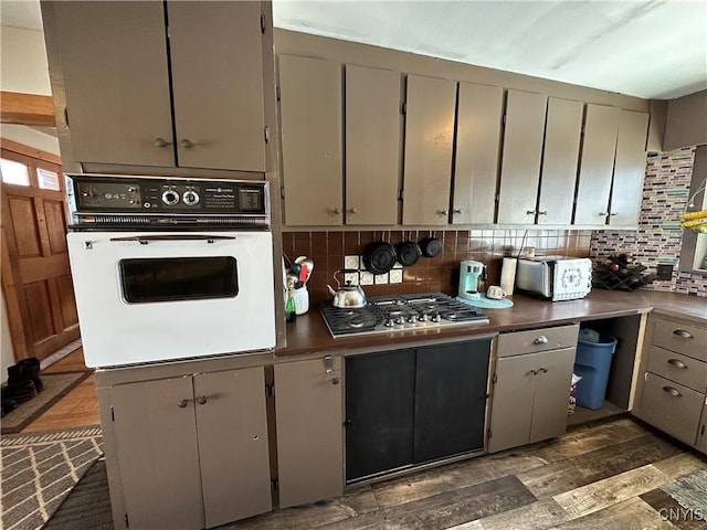 kitchen with dark wood-style floors, stainless steel gas cooktop, gray cabinets, white oven, and backsplash