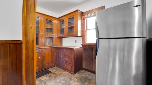 kitchen featuring glass insert cabinets, light countertops, freestanding refrigerator, brown cabinetry, and stone finish floor