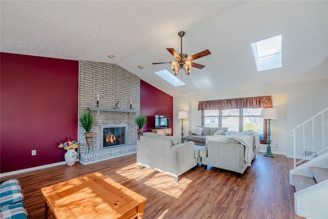 living room featuring a ceiling fan, wood finished floors, vaulted ceiling with skylight, a fireplace, and stairs