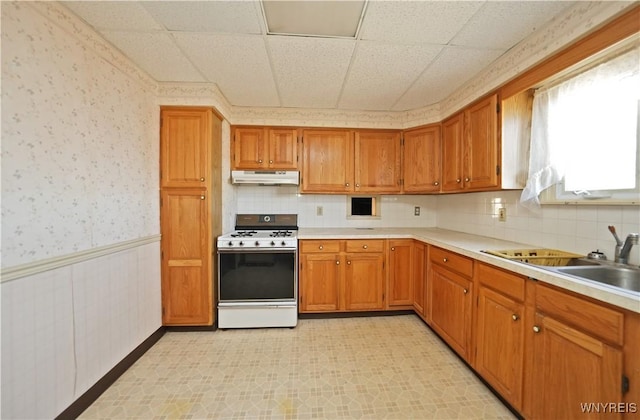 kitchen featuring a wainscoted wall, under cabinet range hood, gas range gas stove, brown cabinetry, and wallpapered walls