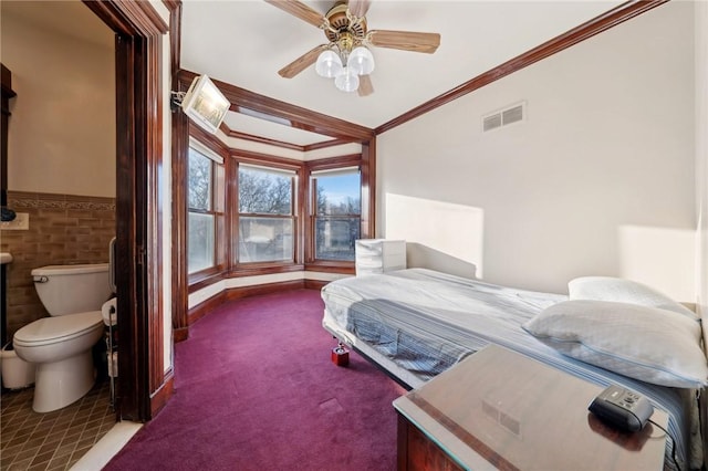 carpeted bedroom with a wainscoted wall, a ceiling fan, visible vents, and ornamental molding