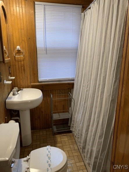 bathroom featuring wood walls, toilet, a shower with shower curtain, brick floor, and a sink