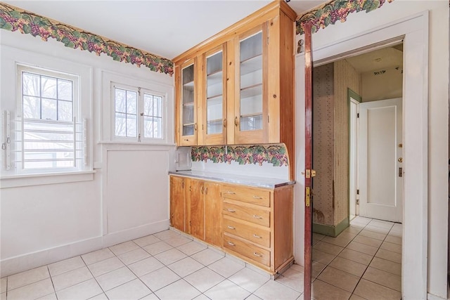 kitchen with baseboards, wallpapered walls, light tile patterned flooring, light countertops, and glass insert cabinets