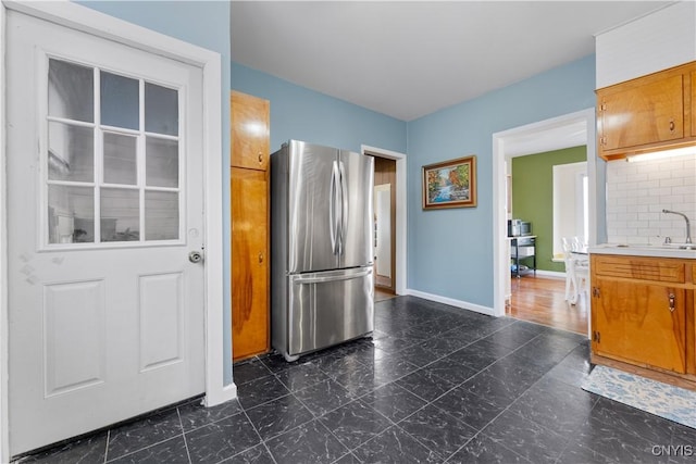 kitchen featuring brown cabinetry, baseboards, freestanding refrigerator, a sink, and tasteful backsplash