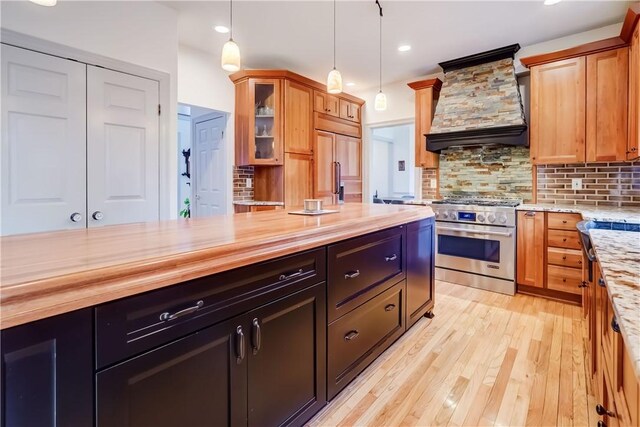 kitchen with butcher block countertops, custom range hood, backsplash, glass insert cabinets, and high end range