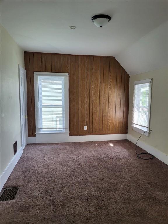 carpeted empty room with lofted ceiling, wooden walls, baseboards, and visible vents