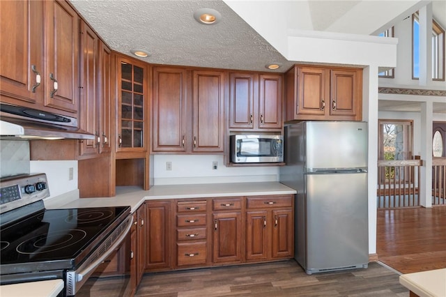 kitchen featuring dark wood-style flooring, light countertops, glass insert cabinets, under cabinet range hood, and appliances with stainless steel finishes