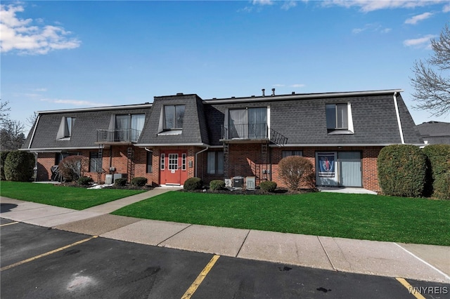 view of front of property with a front lawn, mansard roof, a balcony, roof with shingles, and brick siding