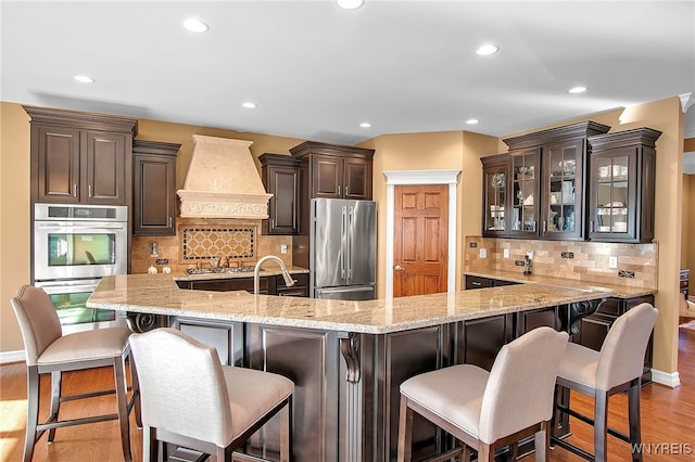 kitchen featuring light wood-style flooring, stainless steel appliances, dark brown cabinetry, a breakfast bar area, and custom exhaust hood