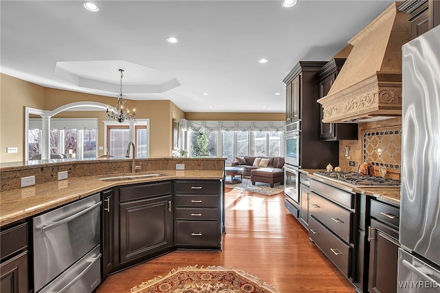 kitchen featuring wood finished floors, custom exhaust hood, stainless steel appliances, a raised ceiling, and a sink