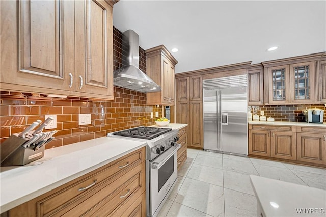 kitchen with tasteful backsplash, glass insert cabinets, light countertops, premium appliances, and wall chimney exhaust hood