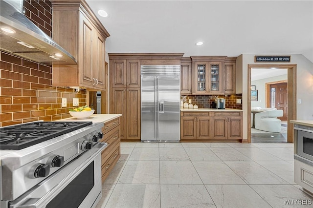 kitchen featuring high end appliances, brown cabinetry, decorative backsplash, light countertops, and wall chimney exhaust hood