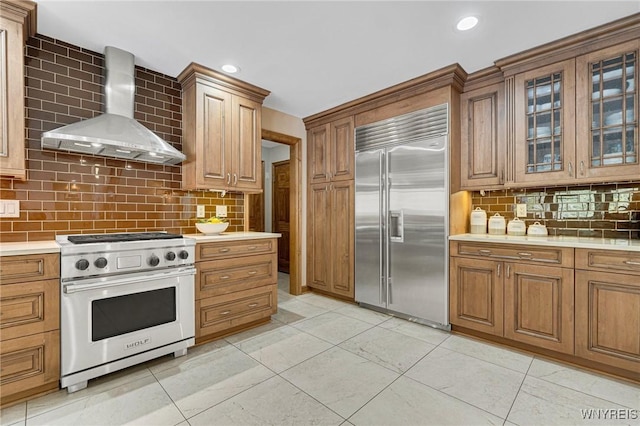 kitchen with light countertops, glass insert cabinets, wall chimney range hood, high quality appliances, and brown cabinets