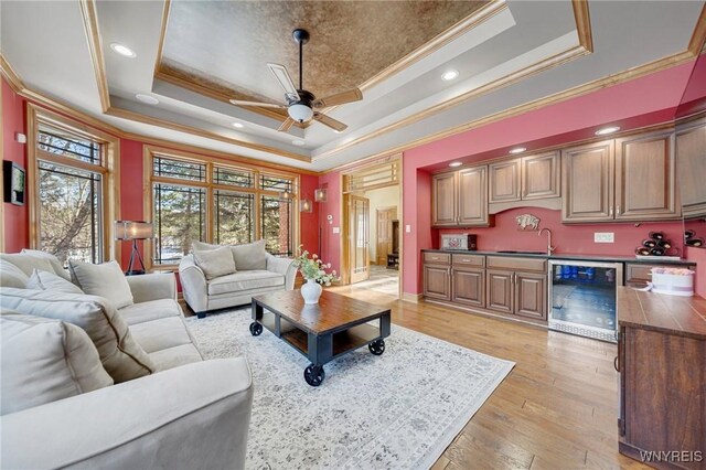 living area with a tray ceiling, wine cooler, light wood-style floors, wet bar, and ceiling fan