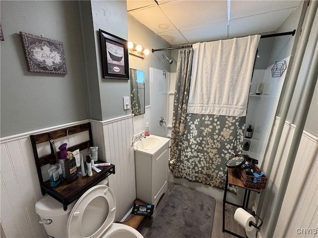 full bathroom featuring shower / bath combo with shower curtain, toilet, a drop ceiling, wainscoting, and vanity