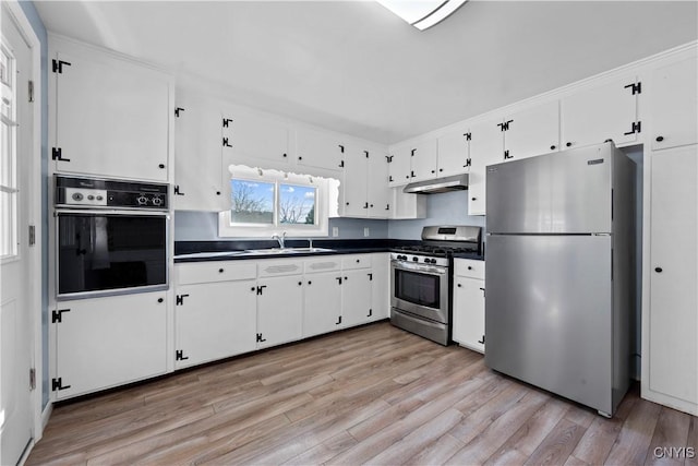 kitchen with a sink, under cabinet range hood, dark countertops, appliances with stainless steel finishes, and white cabinets