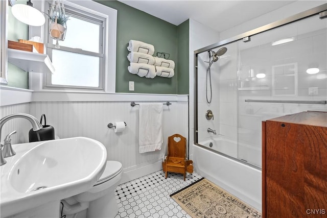 bathroom with a sink, toilet, bath / shower combo with glass door, and wainscoting