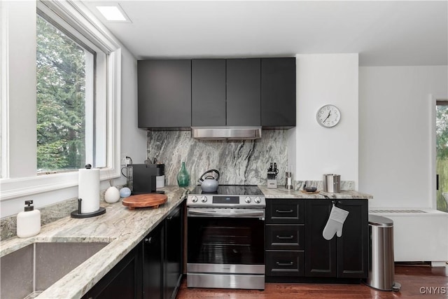 kitchen with light stone countertops, under cabinet range hood, decorative backsplash, stainless steel range with electric cooktop, and dark cabinets