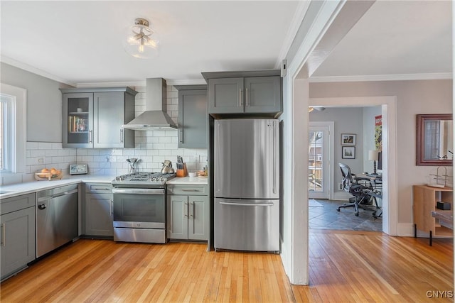 kitchen with gray cabinets, light countertops, appliances with stainless steel finishes, wall chimney range hood, and backsplash