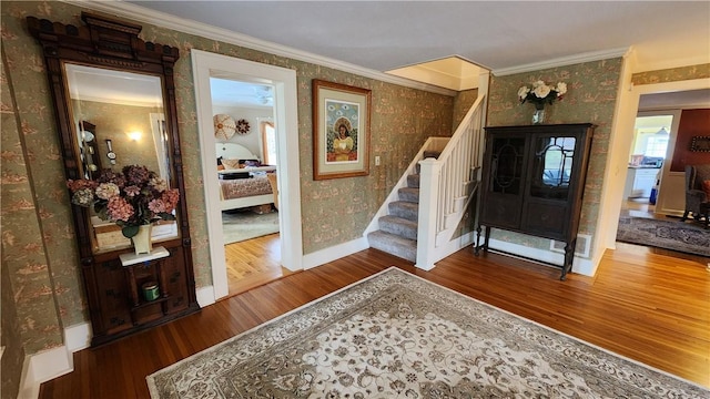 foyer entrance with wallpapered walls, crown molding, baseboards, stairway, and wood finished floors