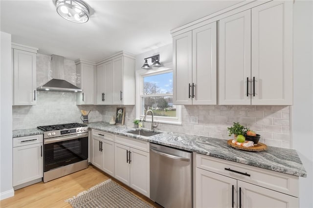 kitchen with a sink, wall chimney range hood, appliances with stainless steel finishes, light wood finished floors, and decorative backsplash