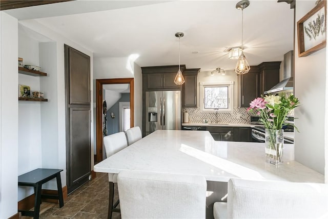 kitchen featuring a peninsula, appliances with stainless steel finishes, a breakfast bar area, wall chimney range hood, and dark brown cabinets