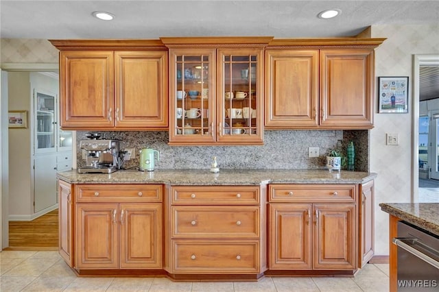 kitchen featuring light stone counters, brown cabinetry, glass insert cabinets, stainless steel dishwasher, and tasteful backsplash