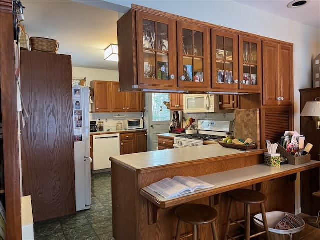 kitchen featuring white appliances, brown cabinetry, a peninsula, glass insert cabinets, and backsplash