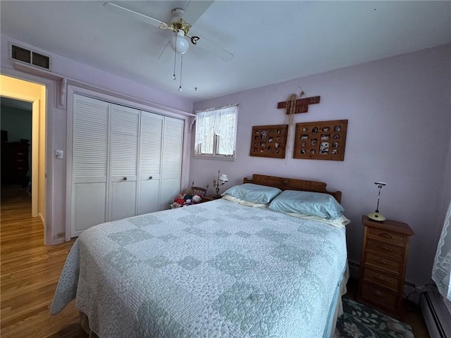 bedroom with wood finished floors, visible vents, ceiling fan, a closet, and a baseboard heating unit