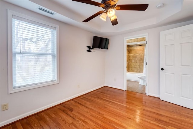 unfurnished bedroom with visible vents, a tray ceiling, connected bathroom, light wood finished floors, and baseboards