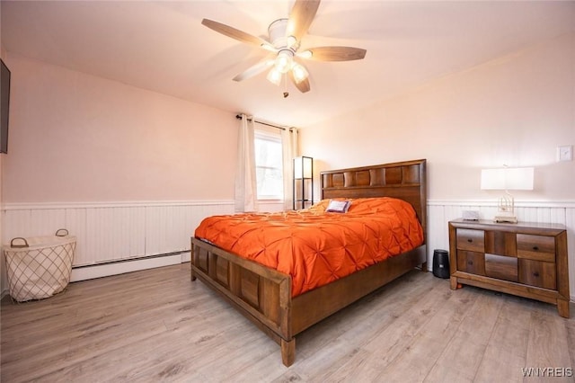 bedroom with baseboard heating, wainscoting, light wood-type flooring, and a ceiling fan