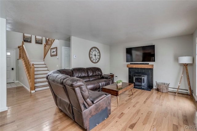 living room featuring a baseboard heating unit, stairway, light wood finished floors, baseboards, and a wood stove