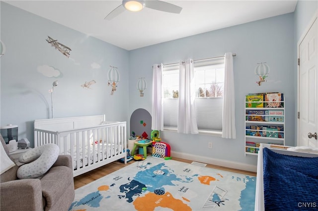 bedroom featuring visible vents, baseboards, a nursery area, wood finished floors, and a ceiling fan