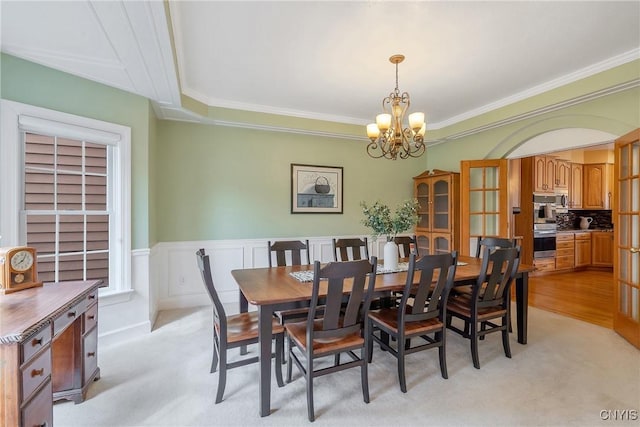 dining space featuring ornamental molding, light carpet, wainscoting, an inviting chandelier, and arched walkways