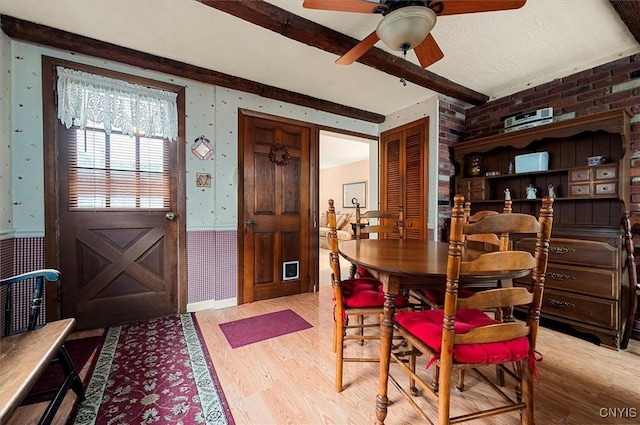 dining room with wood finished floors, a wainscoted wall, wallpapered walls, ceiling fan, and beamed ceiling