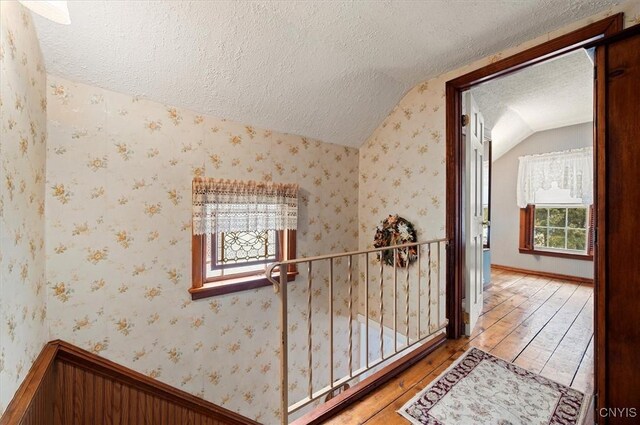 hall featuring light wood finished floors, a textured ceiling, wallpapered walls, and lofted ceiling