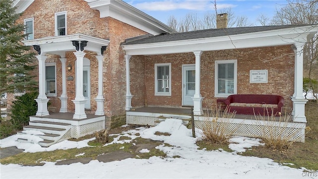 snow covered property entrance featuring a porch, brick siding, and a chimney