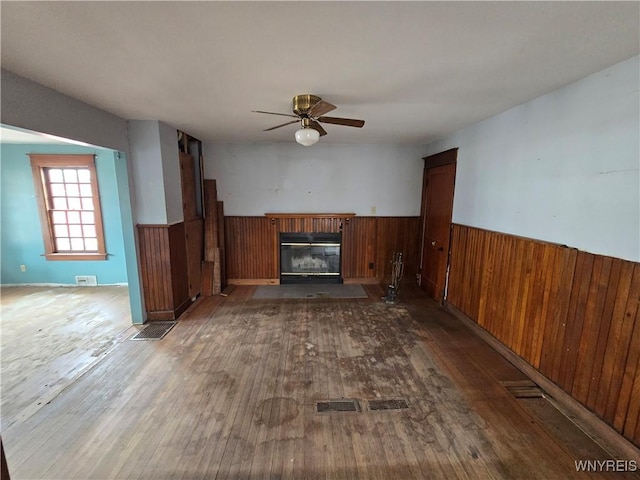 unfurnished living room with wood finished floors, a wainscoted wall, ceiling fan, wood walls, and a glass covered fireplace